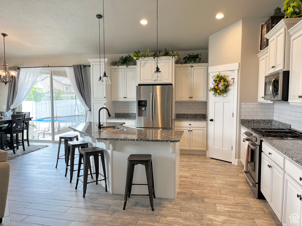 Kitchen featuring dark stone countertops, a center island with sink, appliances with stainless steel finishes, decorative light fixtures, and backsplash