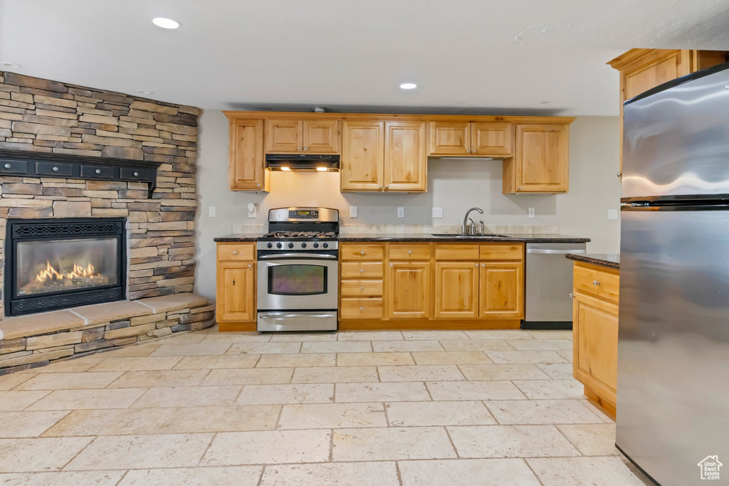 Kitchen with a stone fireplace, stainless steel appliances, light tile flooring, dark stone countertops, and sink