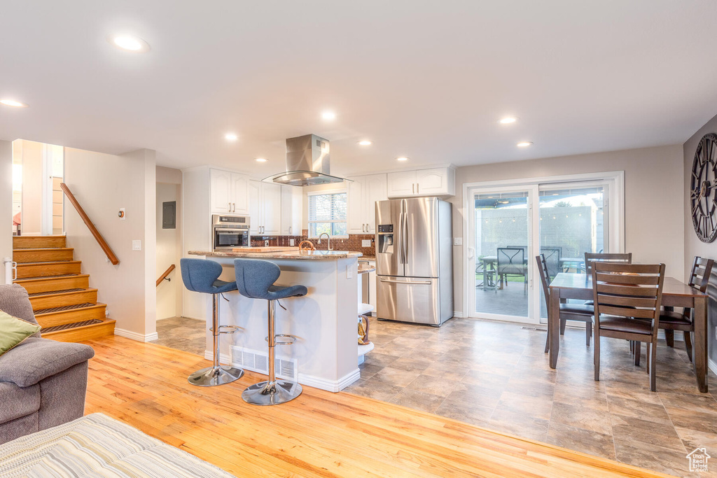 Kitchen featuring light hardwood / wood-style flooring, island range hood, wall oven, stainless steel refrigerator with ice dispenser, and white cabinetry