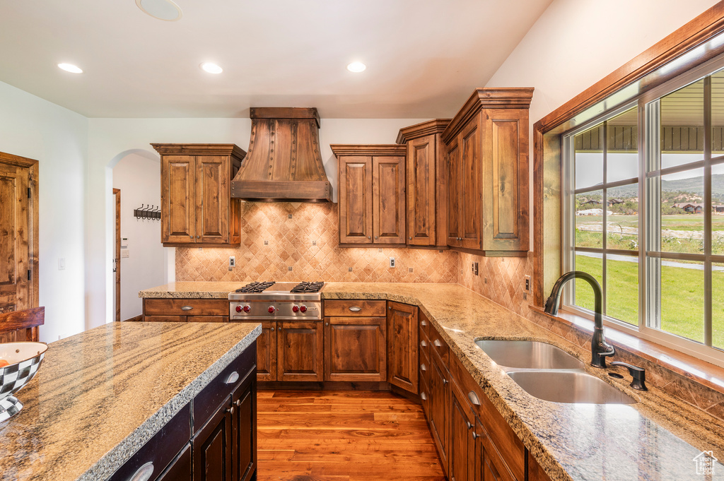 Kitchen with custom exhaust hood, sink, light stone counters, hardwood / wood-style flooring, and tasteful backsplash
