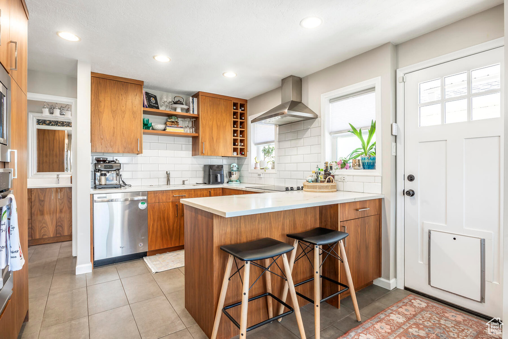 Kitchen with light tile floors, a kitchen bar, dishwasher, wall chimney range hood, and backsplash