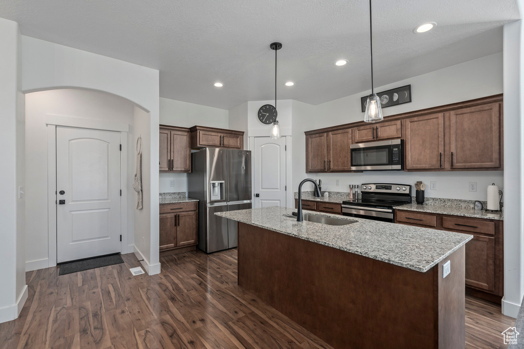 Kitchen with decorative light fixtures, appliances with stainless steel finishes, a kitchen island with sink, dark wood-type flooring, and light stone counters