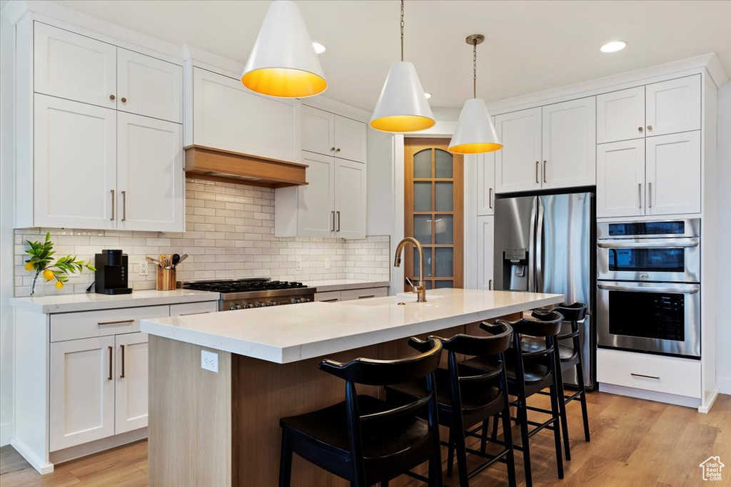 Kitchen with appliances with stainless steel finishes, a kitchen island with sink, light hardwood / wood-style floors, tasteful backsplash, and pendant lighting