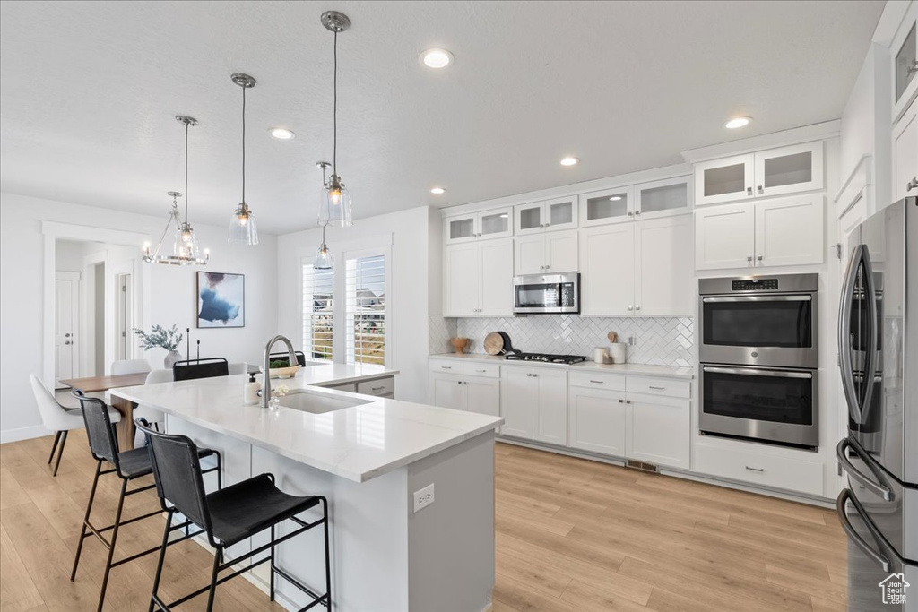 Kitchen with appliances with stainless steel finishes, light hardwood / wood-style flooring, hanging light fixtures, an island with sink, and sink