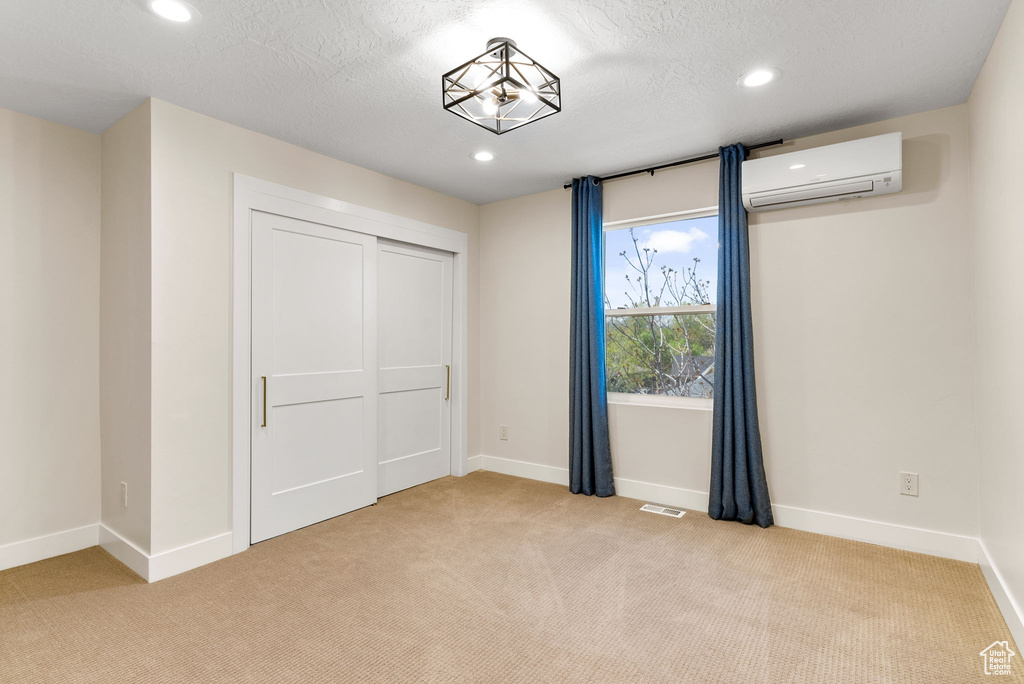 Unfurnished bedroom featuring a closet, a wall mounted air conditioner, light colored carpet, and a textured ceiling