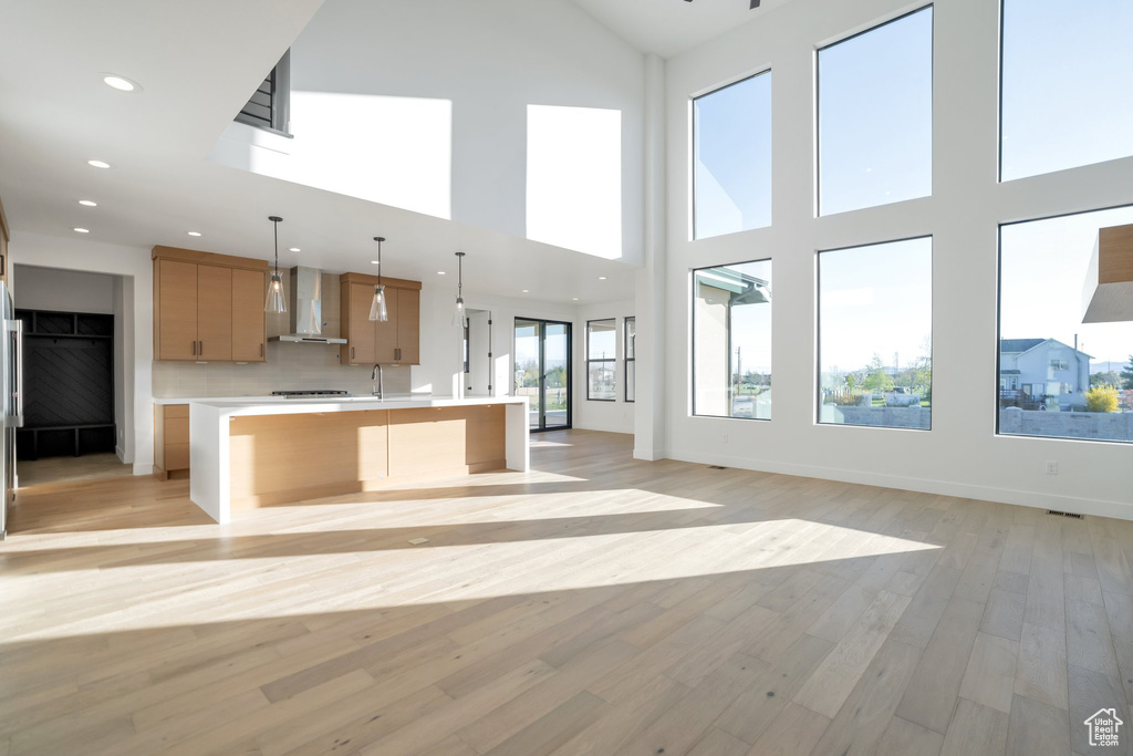 Kitchen with a towering ceiling, a center island with sink, wall chimney range hood, pendant lighting, and light wood-type flooring