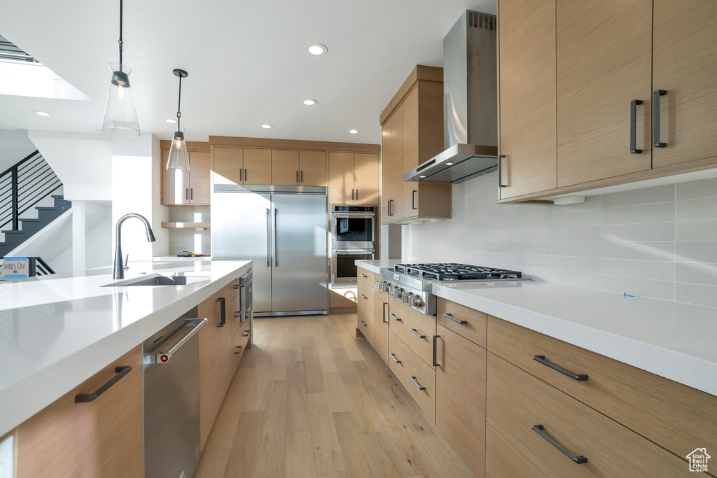 Kitchen featuring wall chimney exhaust hood, decorative light fixtures, light wood-type flooring, appliances with stainless steel finishes, and tasteful backsplash