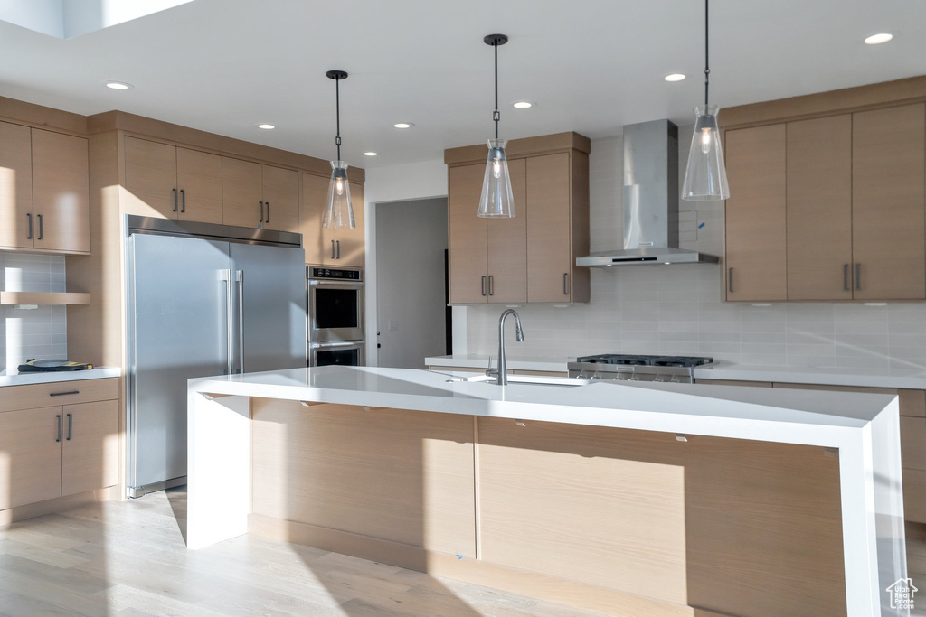 Kitchen featuring wall chimney exhaust hood, hanging light fixtures, light wood-type flooring, backsplash, and an island with sink