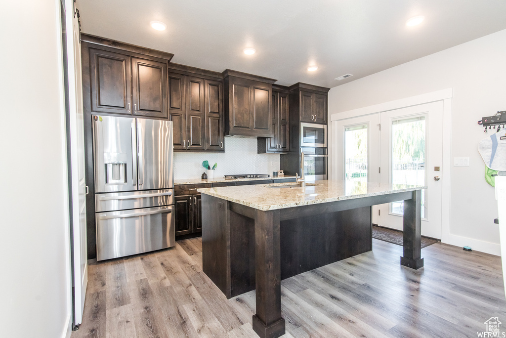Kitchen featuring light stone countertops, appliances with stainless steel finishes, dark brown cabinets, light hardwood / wood-style floors, and an island with sink