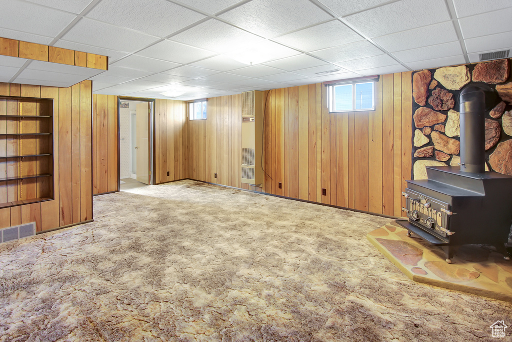 Basement with light colored carpet, a drop ceiling, a wood stove, and wooden walls
