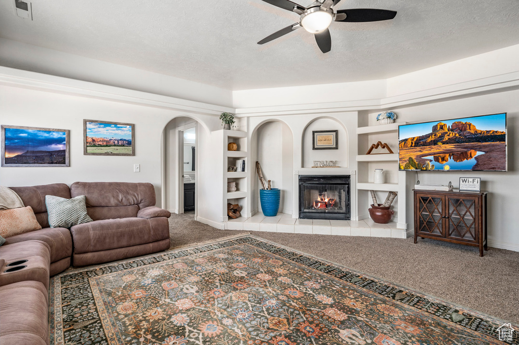 Living room with built in features, ceiling fan, a textured ceiling, a fireplace, and carpet floors