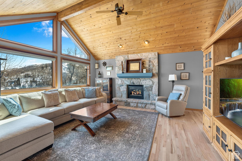 Living room featuring ceiling fan, high vaulted ceiling, light hardwood / wood-style floors, a stone fireplace, and beamed ceiling
