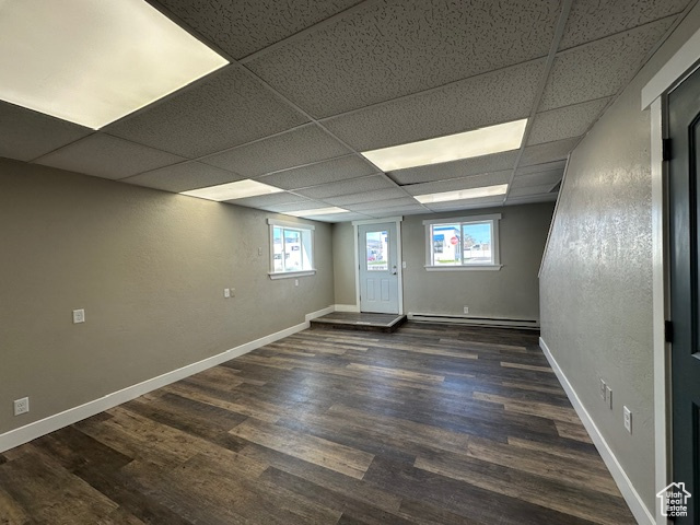 Empty room with a healthy amount of sunlight, dark wood-type flooring, a paneled ceiling, and a baseboard radiator