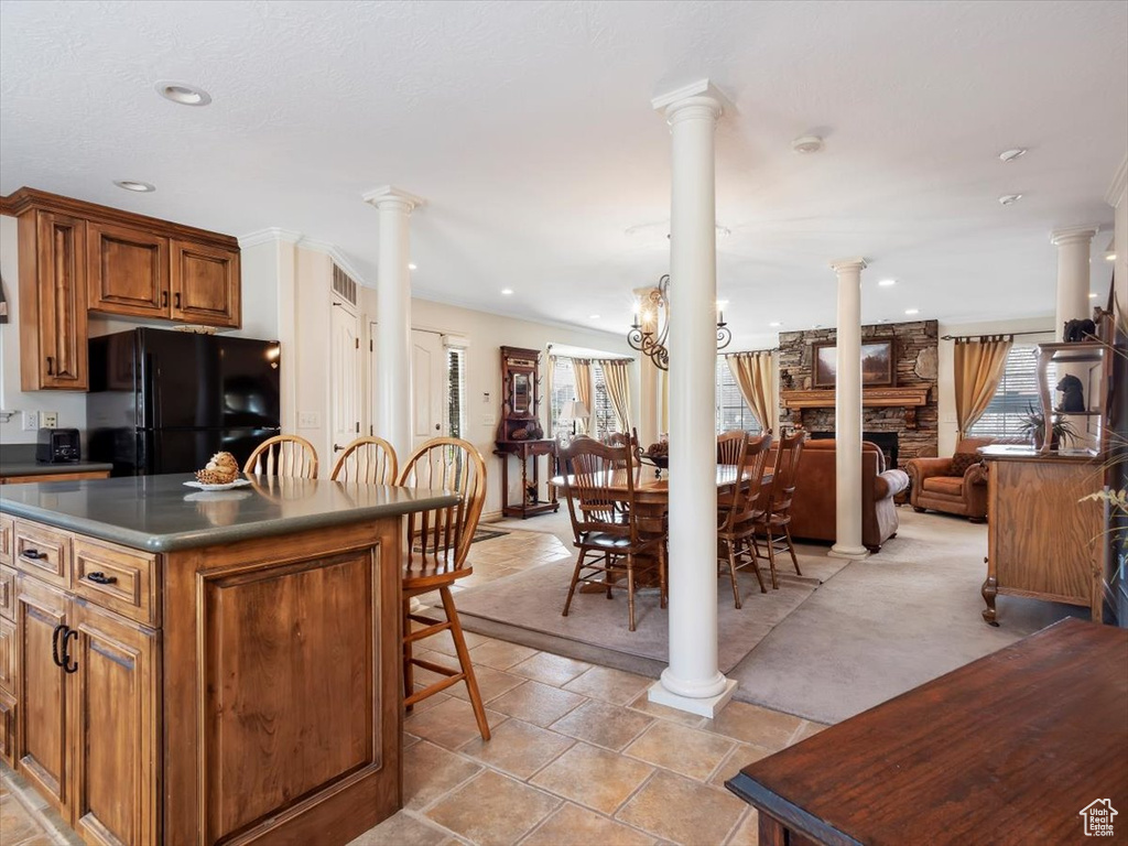 Kitchen featuring light tile floors, an inviting chandelier, a stone fireplace, black refrigerator, and ornate columns