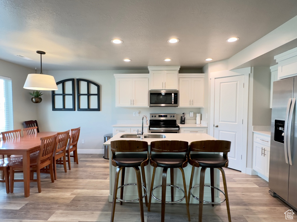 Kitchen featuring light hardwood / wood-style floors, appliances with stainless steel finishes, hanging light fixtures, and a kitchen island with sink