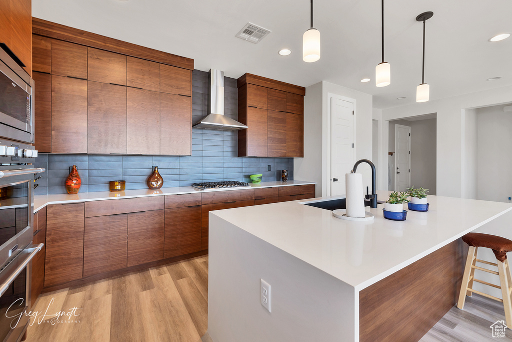 Kitchen featuring light hardwood / wood-style flooring, tasteful backsplash, wall chimney range hood, an island with sink, and sink