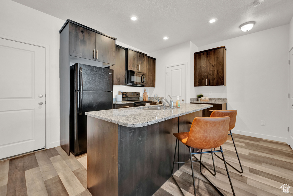 Kitchen featuring black appliances, dark brown cabinets, light stone counters, light hardwood / wood-style floors, and an island with sink