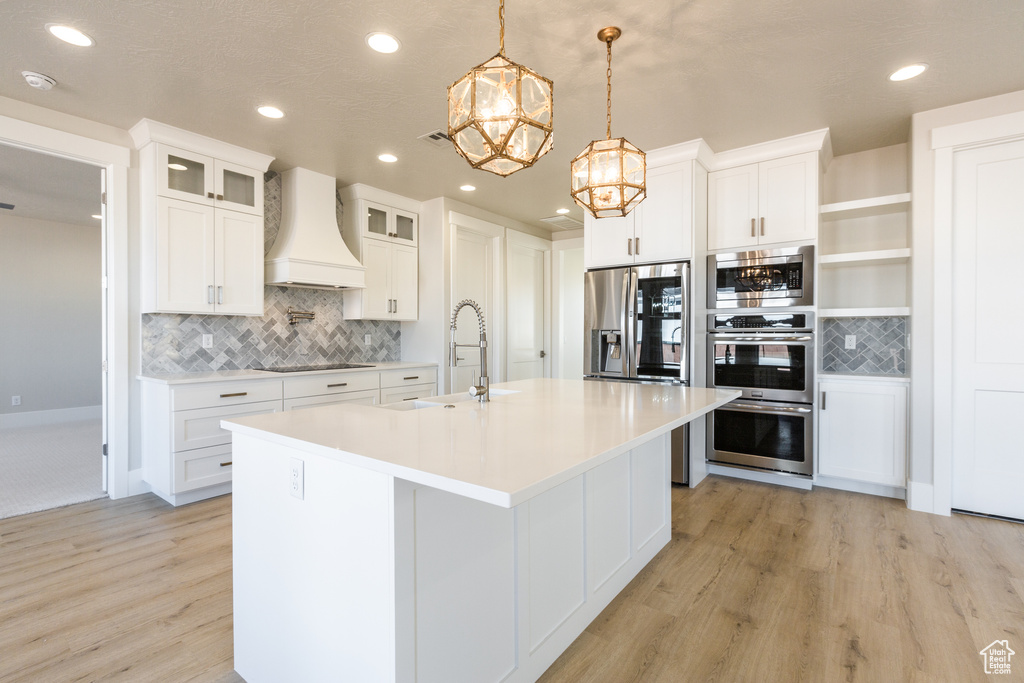 Kitchen featuring backsplash, white cabinetry, light hardwood / wood-style flooring, custom range hood, and a kitchen island with sink