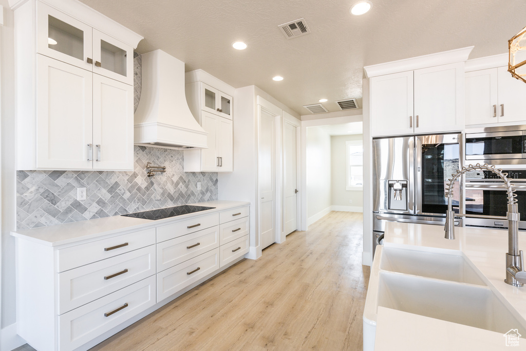 Kitchen with custom exhaust hood, light wood-type flooring, white cabinetry, backsplash, and appliances with stainless steel finishes