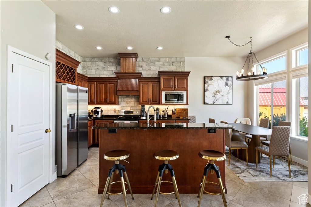 Kitchen featuring appliances with stainless steel finishes, backsplash, dark stone counters, a kitchen island with sink, and light tile floors