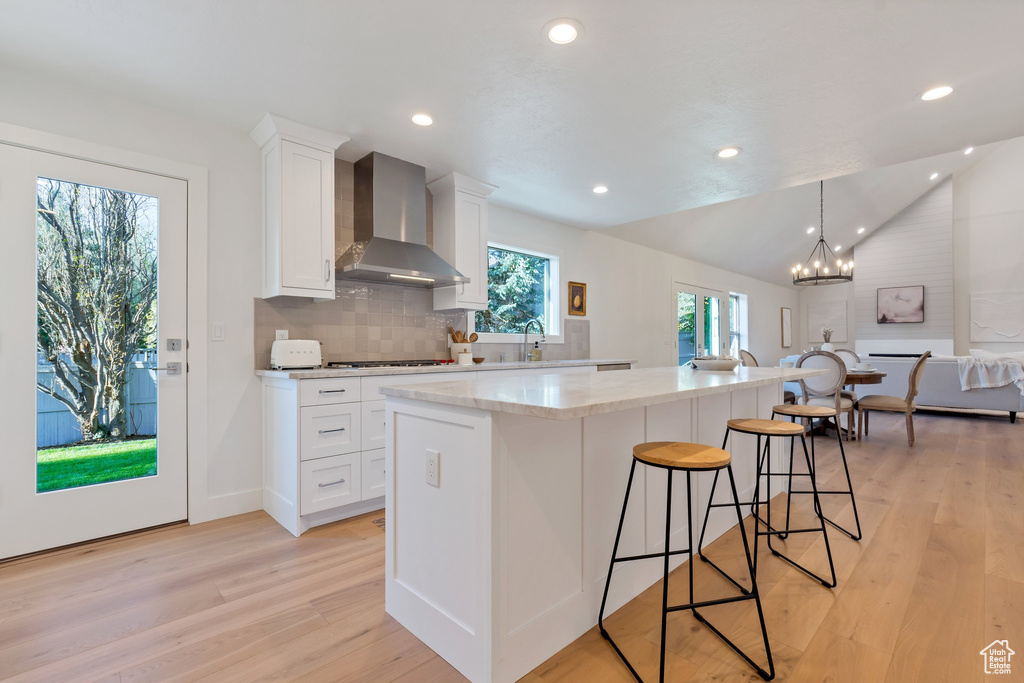 Kitchen featuring hanging light fixtures, light hardwood / wood-style floors, an island with sink, wall chimney exhaust hood, and white cabinets