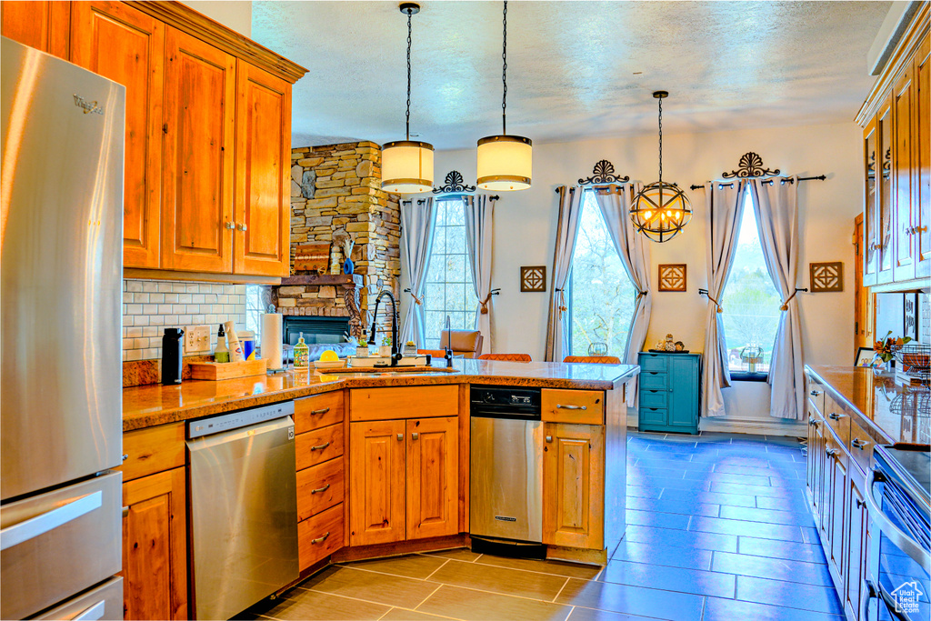 Kitchen with a stone fireplace, appliances with stainless steel finishes, backsplash, decorative light fixtures, and sink