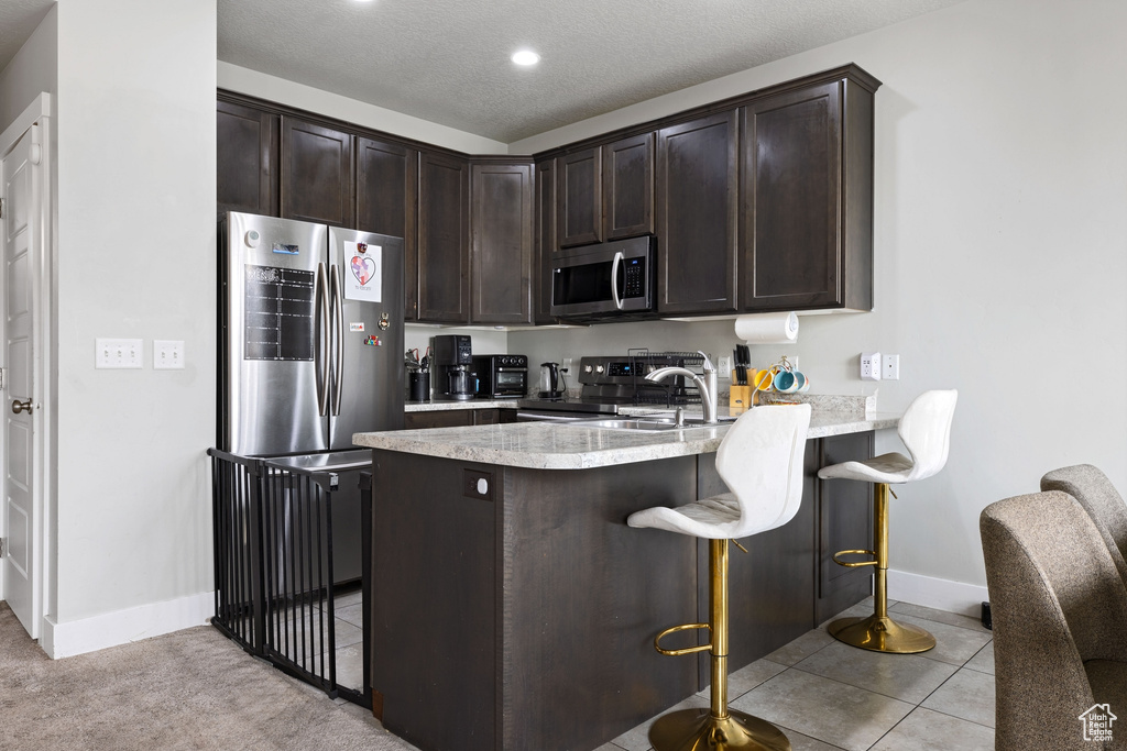 Kitchen featuring dark brown cabinets, kitchen peninsula, appliances with stainless steel finishes, light carpet, and a breakfast bar