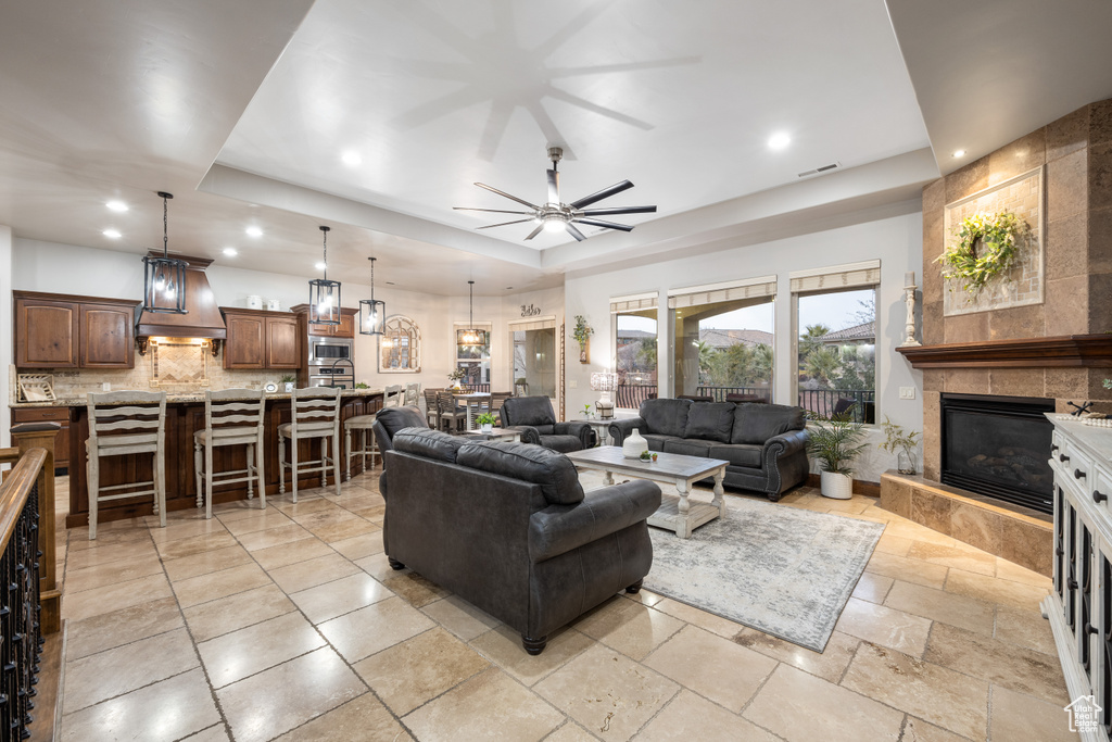 Living room featuring ceiling fan, light tile floors, a tray ceiling, and a tiled fireplace