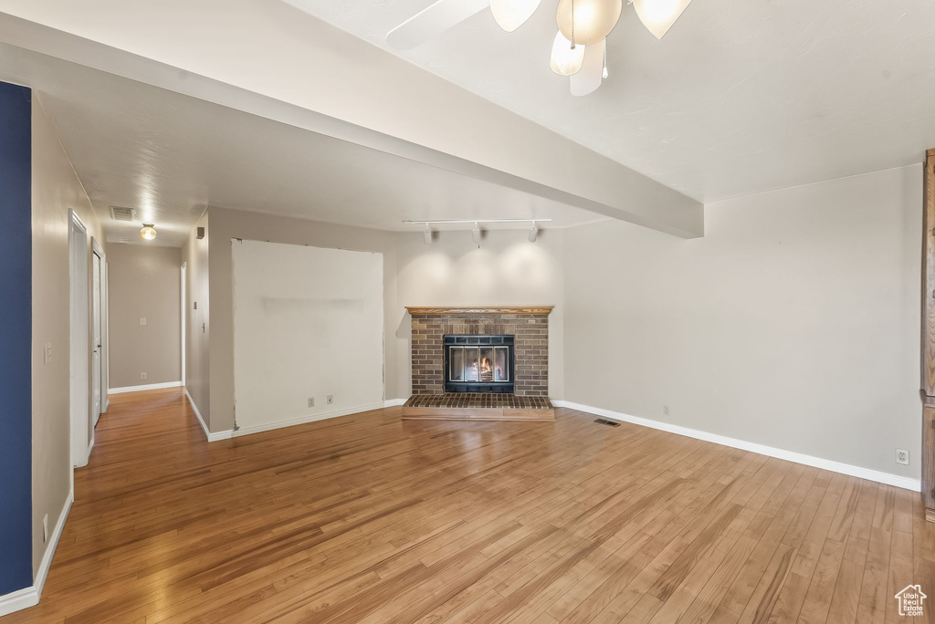 Unfurnished living room with a brick fireplace, ceiling fan, track lighting, beam ceiling, and light wood-type flooring