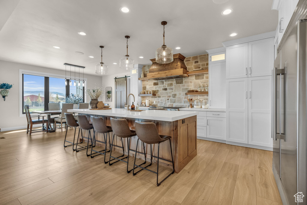 Kitchen with premium range hood, stainless steel built in fridge, light hardwood / wood-style floors, an island with sink, and white cabinets