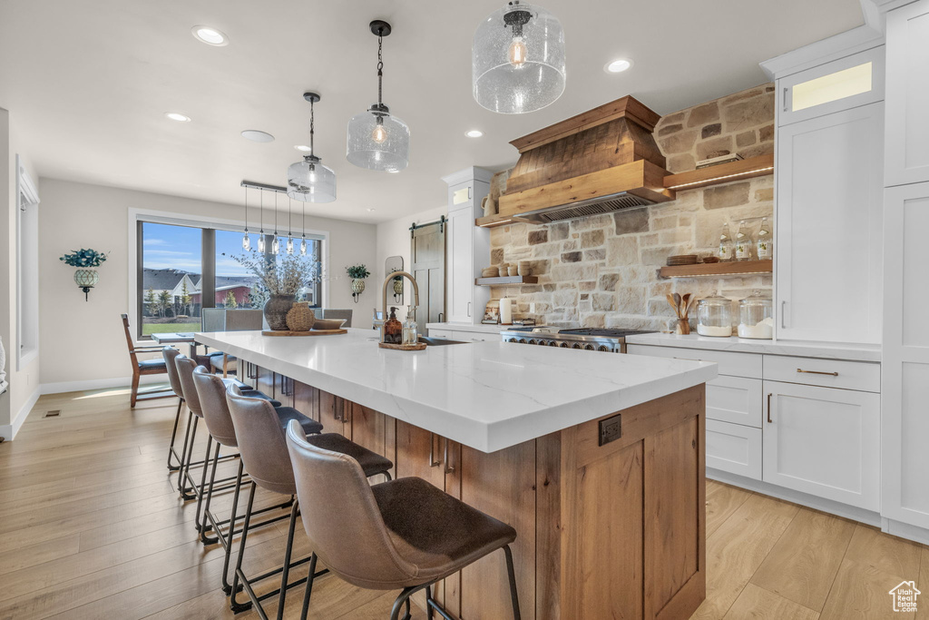 Kitchen with light stone countertops, premium range hood, an island with sink, sink, and light wood-type flooring