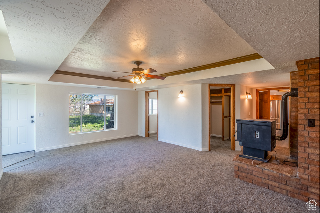Unfurnished living room featuring carpet floors, ceiling fan, brick wall, a tray ceiling, and a wood stove