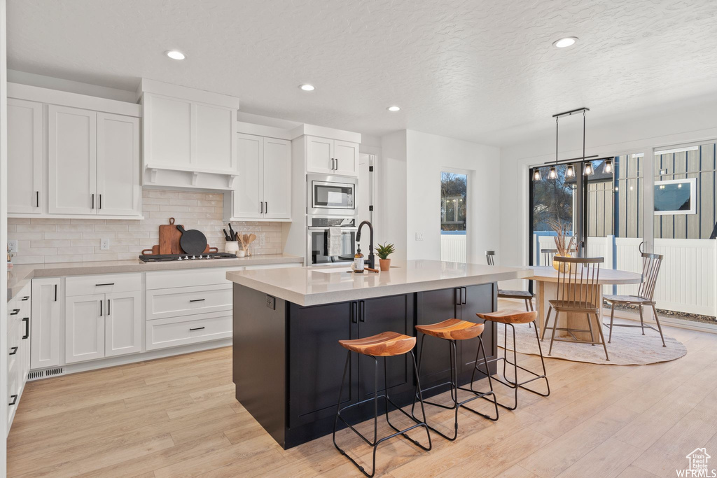 Kitchen featuring white cabinets, stainless steel appliances, light wood-type flooring, decorative light fixtures, and an island with sink