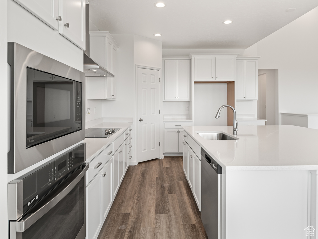 Kitchen with appliances with stainless steel finishes, white cabinets, wall chimney range hood, dark hardwood / wood-style flooring, and a center island with sink