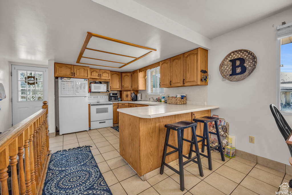 Kitchen featuring white appliances, light tile flooring, kitchen peninsula, sink, and a kitchen bar