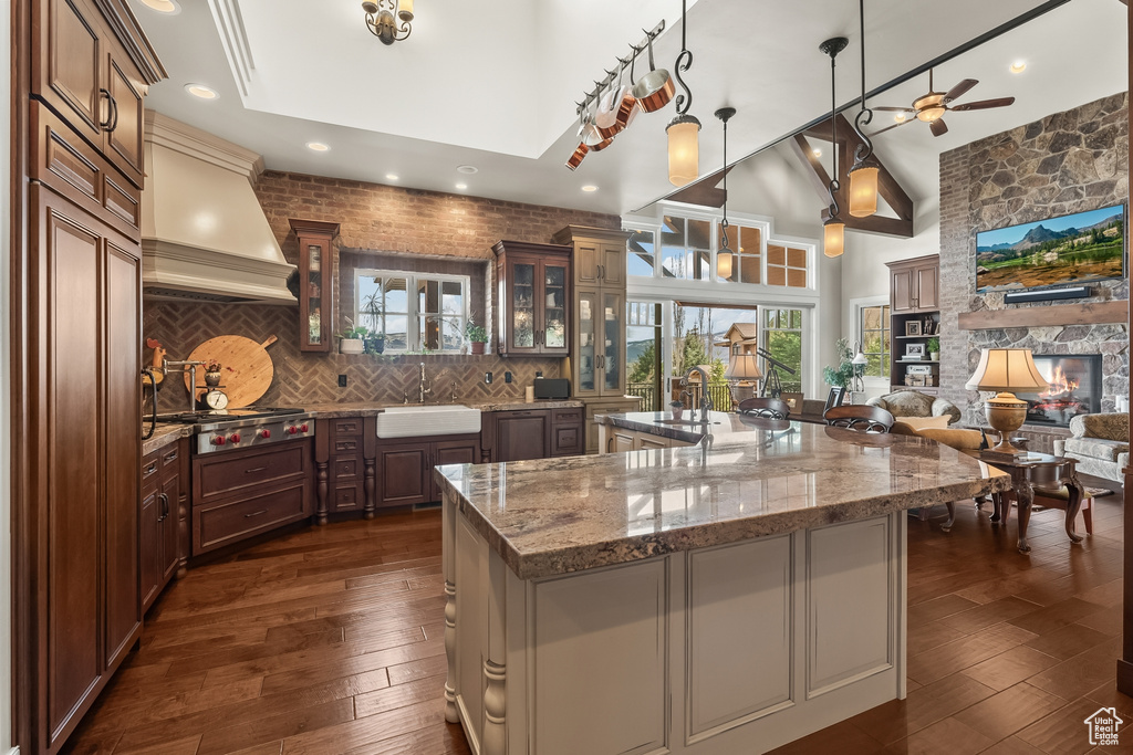 Kitchen featuring premium range hood, tasteful backsplash, a large island with sink, a stone fireplace, and ceiling fan