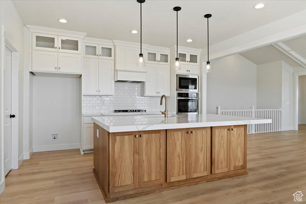 Kitchen with appliances with stainless steel finishes, tasteful backsplash, a kitchen island with sink, pendant lighting, and light wood-type flooring