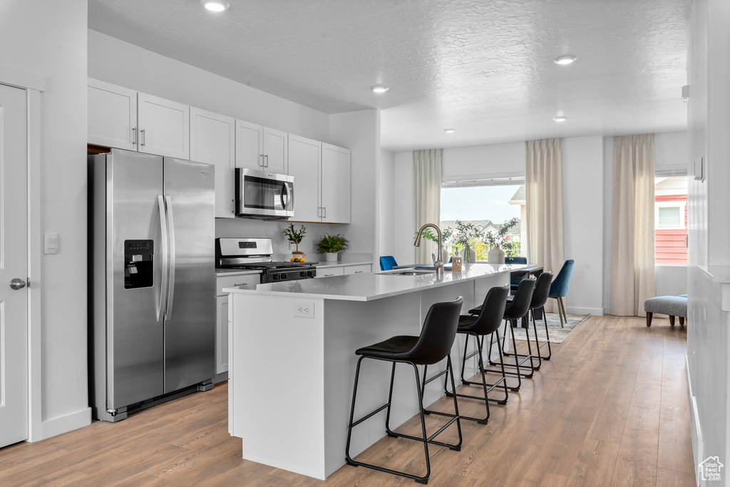 Kitchen with light wood-type flooring, white cabinetry, stainless steel appliances, sink, and a center island with sink