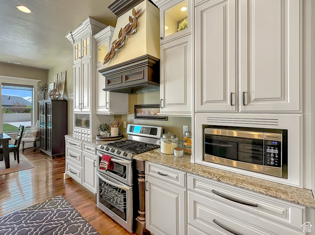 Kitchen with premium range hood, light stone countertops, light hardwood / wood-style flooring, white cabinetry, and appliances with stainless steel finishes