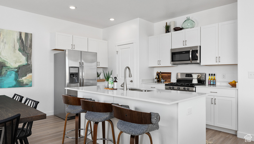 Kitchen featuring an island with sink, white cabinets, sink, stainless steel appliances, and light hardwood / wood-style flooring