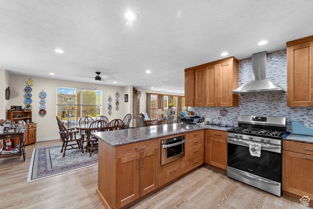 Kitchen with kitchen peninsula, appliances with stainless steel finishes, wall chimney range hood, tasteful backsplash, and light hardwood / wood-style floors