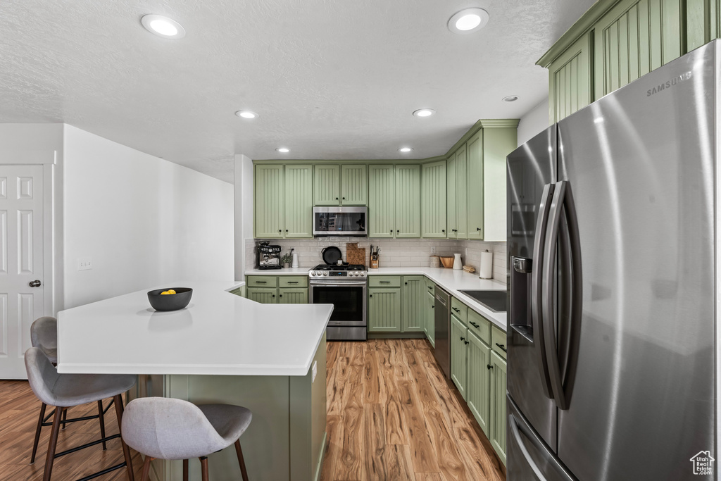 Kitchen with appliances with stainless steel finishes, green cabinets, light hardwood / wood-style floors, tasteful backsplash, and a kitchen breakfast bar