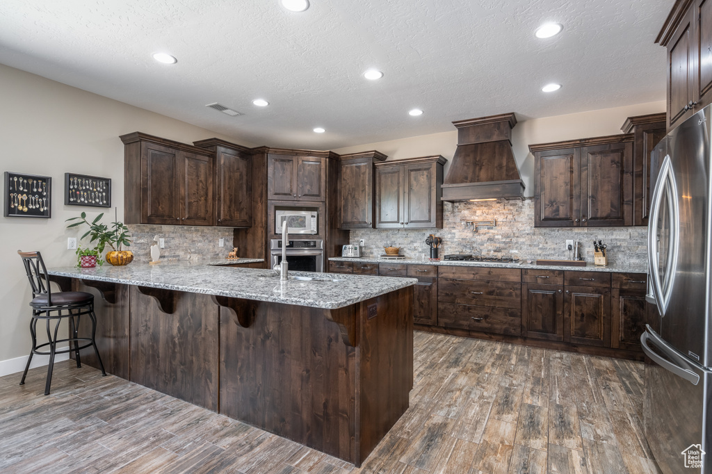 Kitchen with appliances with stainless steel finishes, tasteful backsplash, hardwood / wood-style flooring, premium range hood, and light stone countertops