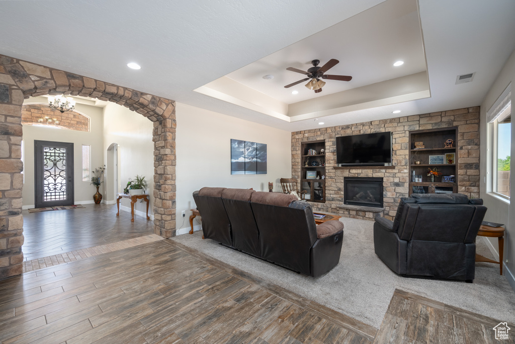 Living room featuring a stone fireplace, carpet flooring, ceiling fan, and a tray ceiling