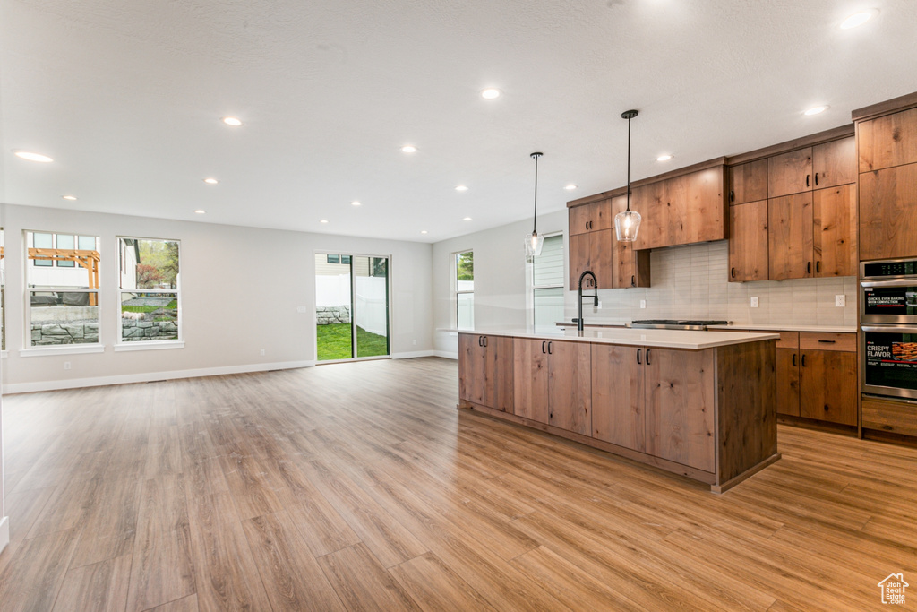 Kitchen featuring appliances with stainless steel finishes, tasteful backsplash, light wood-type flooring, a center island with sink, and pendant lighting