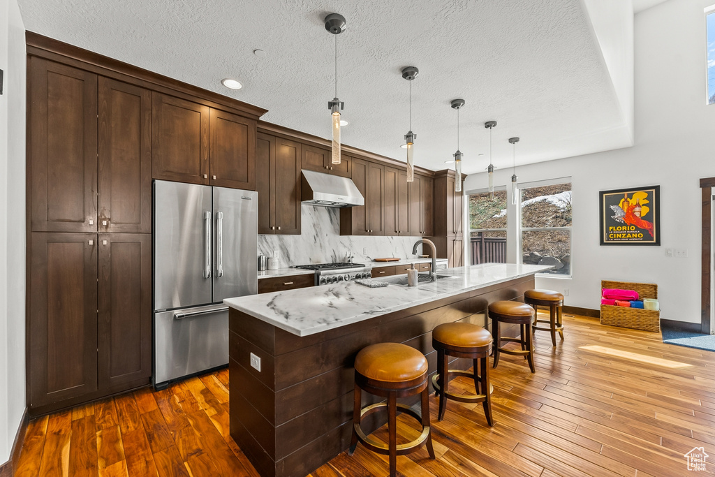 Kitchen featuring light stone countertops, high end fridge, decorative light fixtures, dark hardwood / wood-style floors, and an island with sink