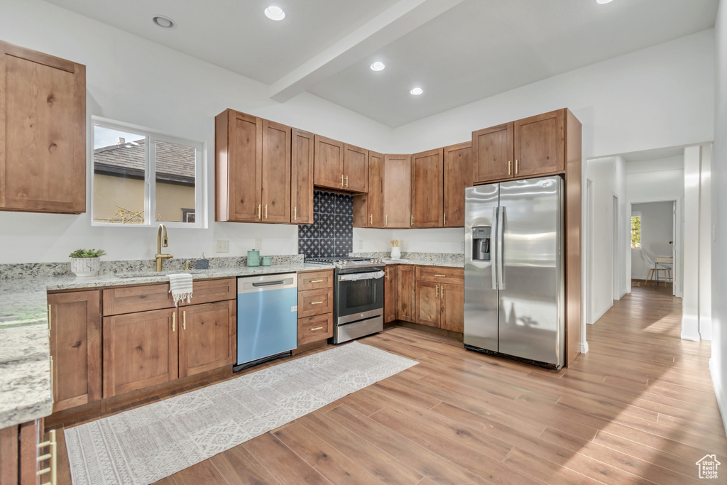 Kitchen with beamed ceiling, appliances with stainless steel finishes, sink, light stone counters, and light hardwood / wood-style floors