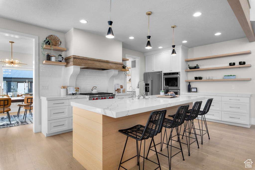 Kitchen with appliances with stainless steel finishes, white cabinets, backsplash, light wood-type flooring, and a center island with sink