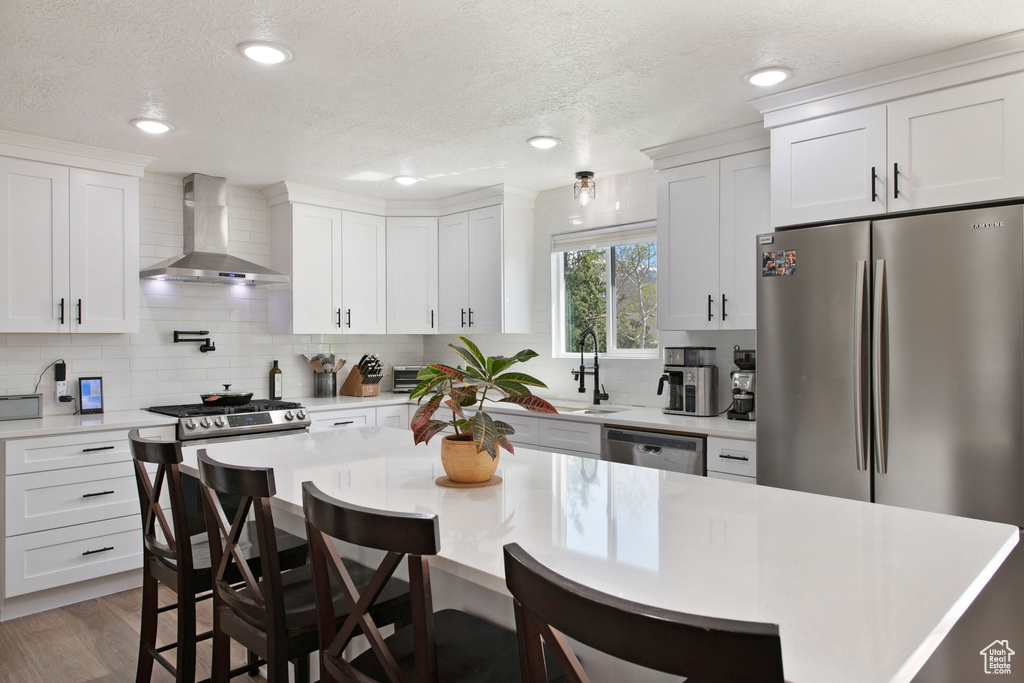 Kitchen with white cabinets, wall chimney range hood, stainless steel appliances, hardwood / wood-style flooring, and tasteful backsplash