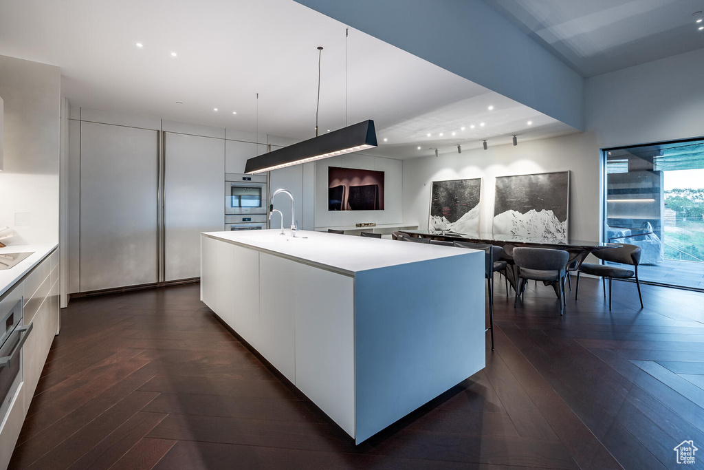 Kitchen with sink, dark parquet flooring, stainless steel double oven, an island with sink, and pendant lighting
